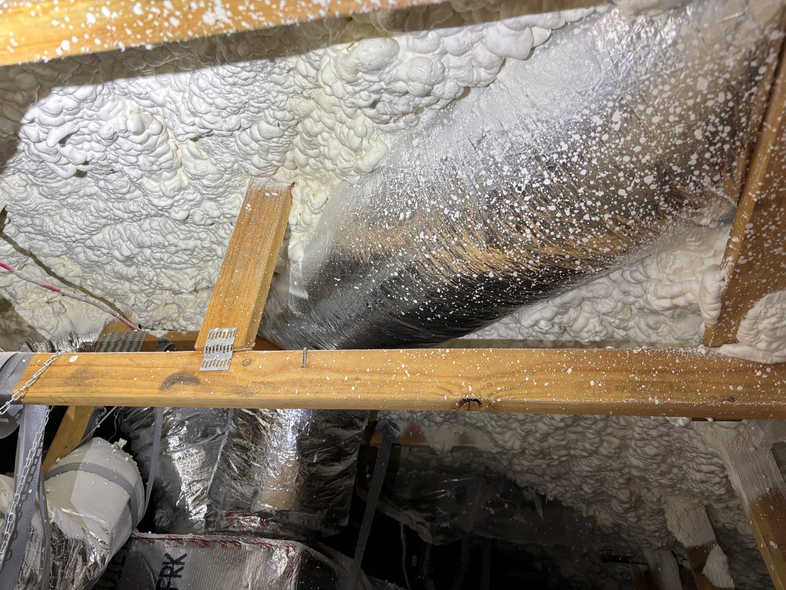 Attic with spray foam covering the roof deck and oversprayed onto a foil-wrapped flex duct, joists, and a wood beam.